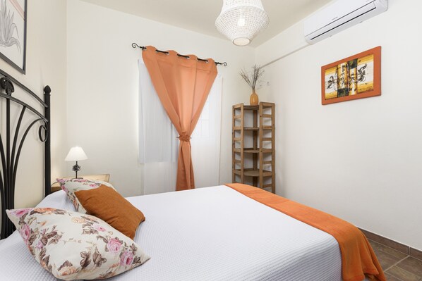Una camera d'albergo con un letto, un comodino, una lampada, una finestra con tende, uno scaffale in legno e un'opera d'arte appesa al muro.