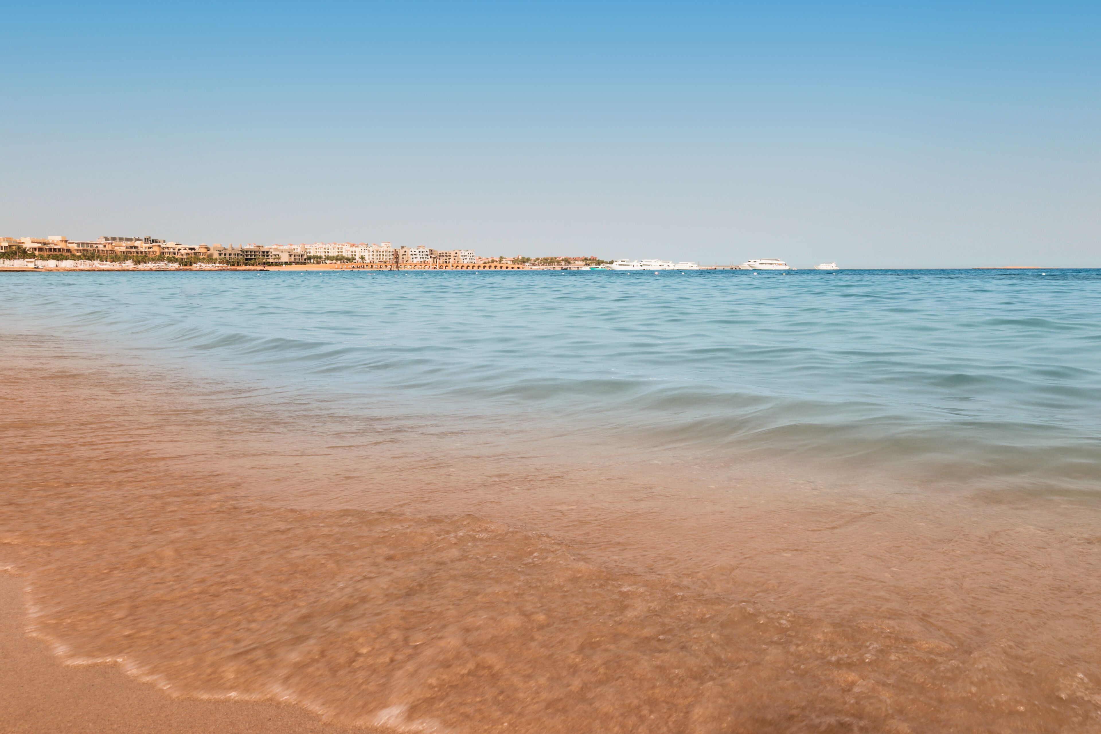 A beach with clear water, a sandy shore, and a distant cityscape.
