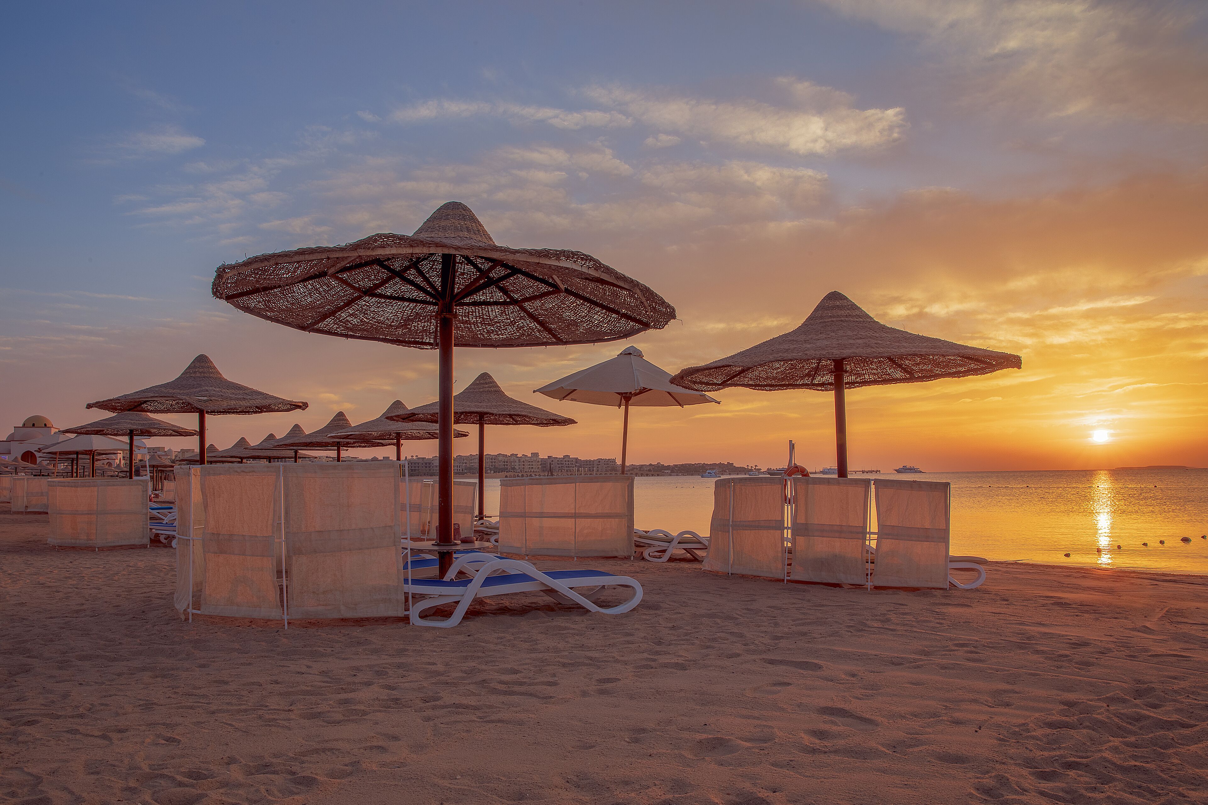 A beach with sun loungers and thatched umbrellas, a calm sea, and a sunset.