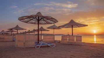 A beach with sun loungers and thatched umbrellas, a calm sea, and a sunset.