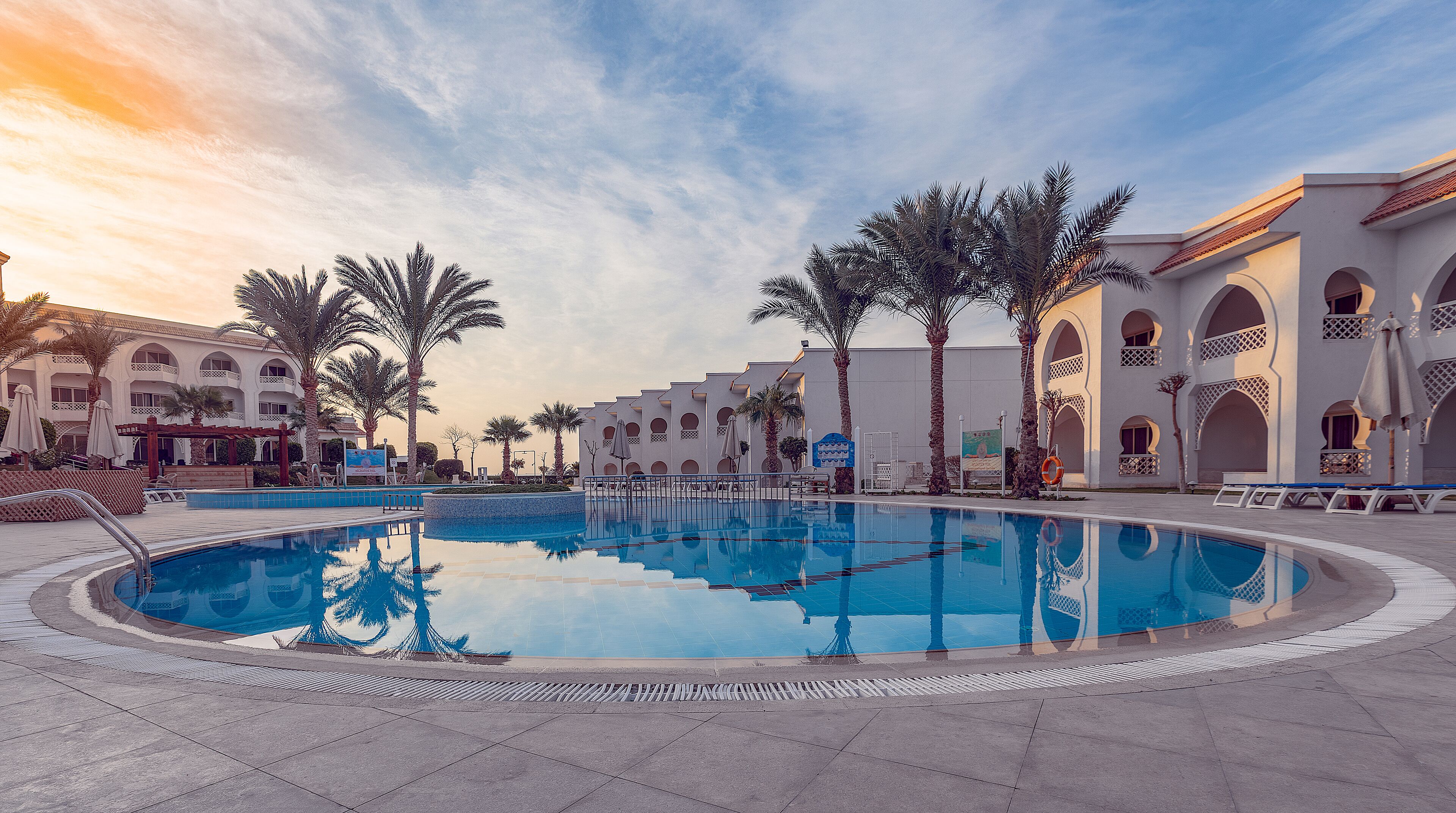 A swimming pool surrounded by palm trees and a building with arched windows.