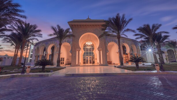 A grand building with arched entrances and a central dome, surrounded by palm trees and a paved walkway. - Old Palace Sahl Hasheseh (Sahl Hasheeh)