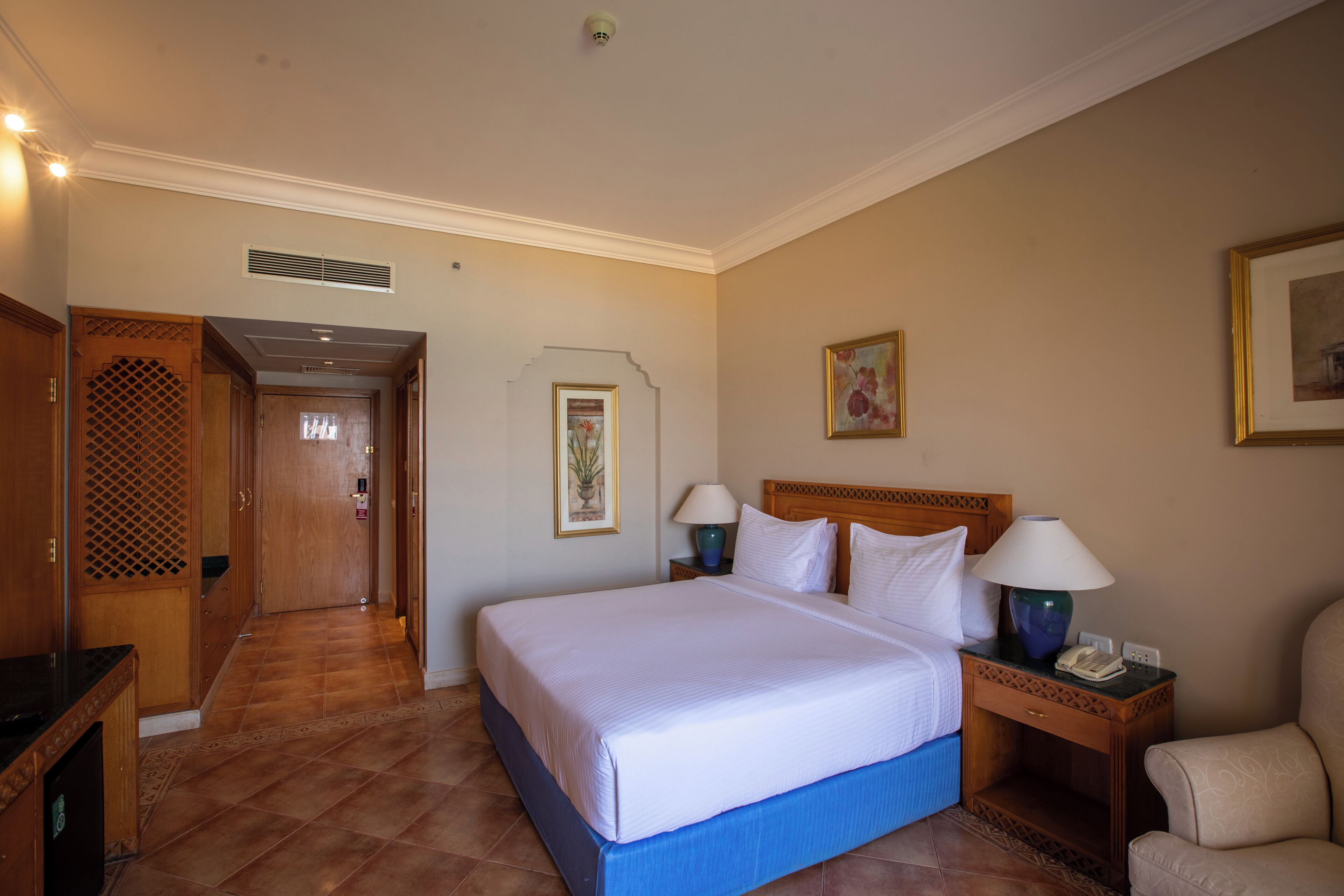 A hotel room with a large bed, two bedside tables with lamps, a wooden headboard, a door with a decorative panel, and a framed picture on the wall.