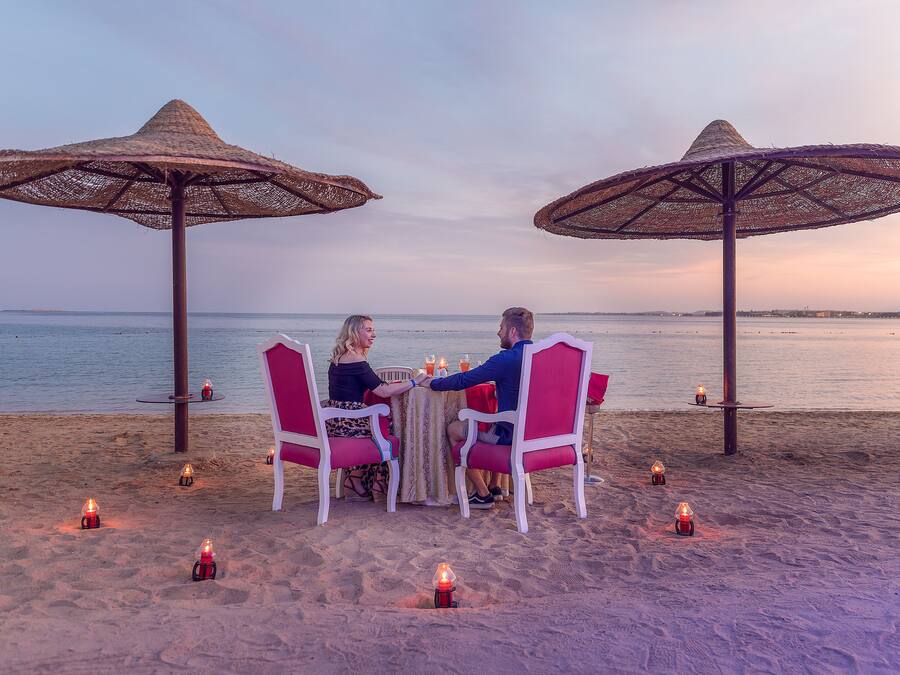 A couple dining at a beachside table with two large umbrellas, surrounded by candles.