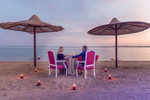 A couple dining at a beachside table with two large umbrellas, surrounded by candles. - Old Palace Sahl Hasheseh (Sahl Hasheeh)