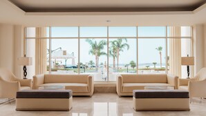 A spacious room with two beige sofas, a glass coffee table, and a large window offering a view of palm trees and a beach. - Old Palace Sahl Hasheseh (Sahl Hasheeh)
