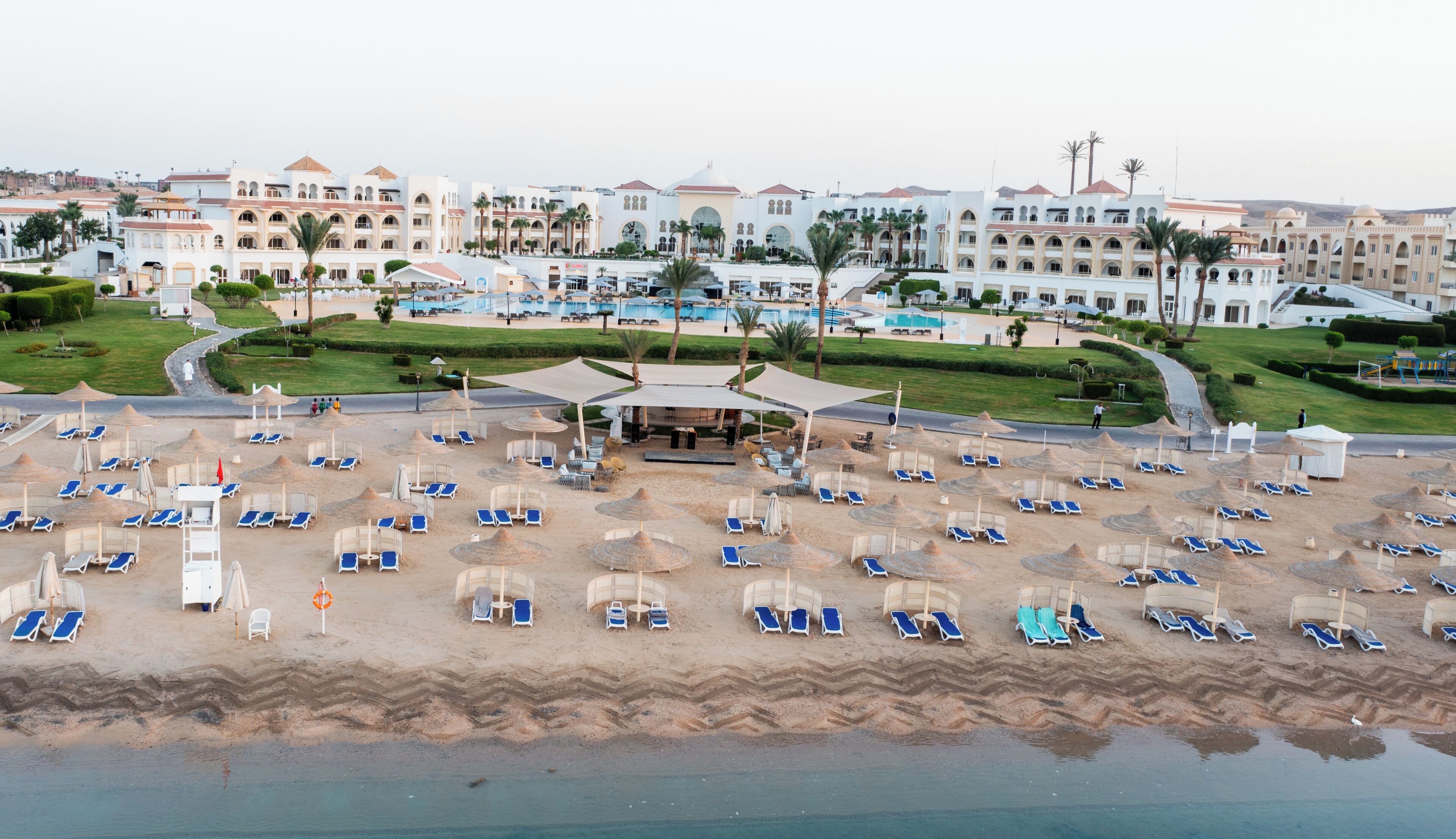 Ein Resort mit großzügigem Poolbereich, zahlreichen Liegestühlen und einem Strand mit Sonnenschirmen.