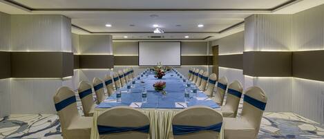 Un salón de conferencias con una larga mesa, sillas color beige adornadas con cintas azules y una pantalla de proyector al frente.