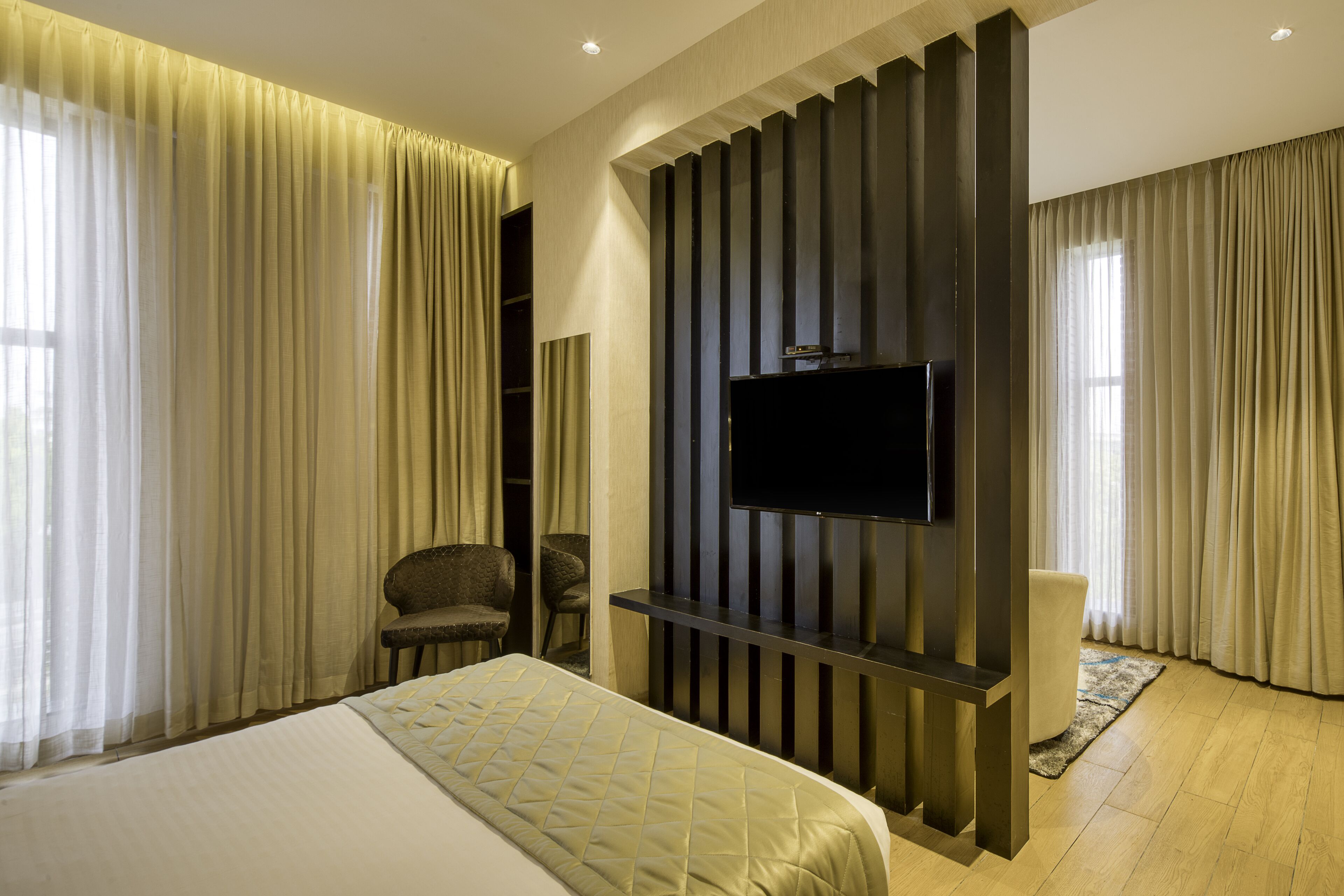 A hotel room with a large bed, a flat-screen TV mounted on a dark panel, a seating area with two chairs, and a large window with curtains.
