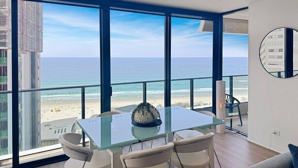 A modern dining area with a glass table and chairs, overlooking a beach and ocean.