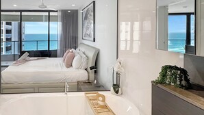 A modern bedroom with a large bed, a bathtub, and a view of the ocean.
