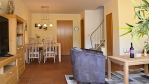 A modern living room with a sofa, a dining area with chairs and a table, a TV, and a plant. - Dirks Ferienhaus Fleute - Holiday home Fleute (Varel)