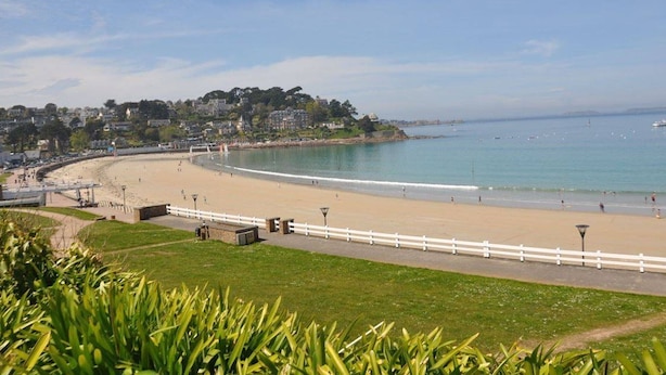 A coastal beach with a white fence, a paved walkway, and a view of the sea and buildings in the background.