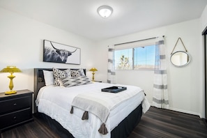 A neatly arranged bedroom with a large bed, two bedside tables with lamps, a window with curtains, and a wall-mounted mirror.