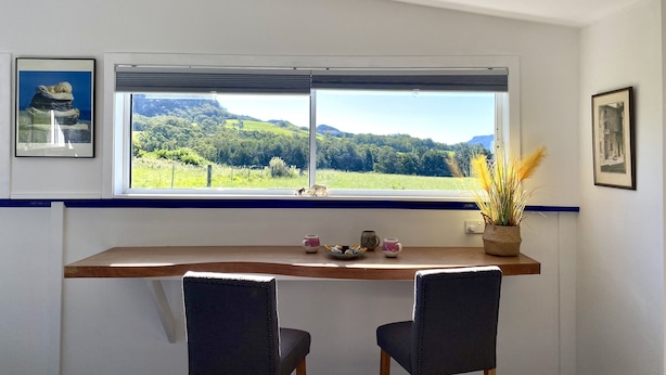A dining area with a wooden table and two chairs, a window with a view of green hills, and two framed pictures on the wall. - Casa Azure - quaint studio with a mediterranean touch (Kangaroo Valley)