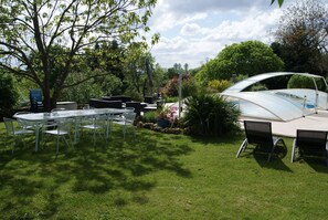An outdoor patio with a glass table and chairs, surrounded by lush greenery and a swimming pool.