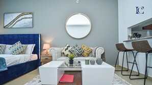 A modern bedroom with a blue headboard, a round mirror, a white sofa with patterned cushions, a small table with a red book, and a kitchen area with bar stools. - Lake View Brand New Studio in X1 Tower JLT - LND (Dubai)