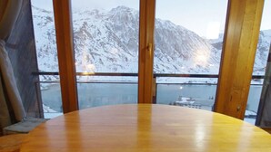 A wooden table in a room with large windows offering a view of snow-covered mountains and a body of water. - Tignes: Classic Apartment with Lake View, 5 Sleeps (Tignes)