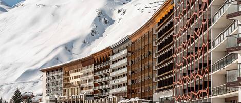 A hotel with multiple balconies, a ski slope in the background, and snow-covered surroundings.