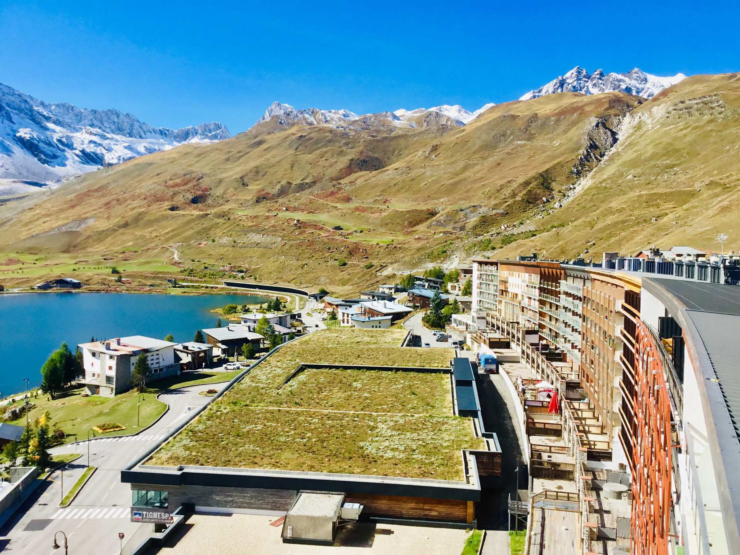 A modern hotel with a rooftop garden, situated by a lake with mountains in the background.