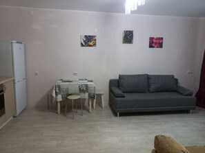 A living room with a grey sofa, a small table with chairs, a refrigerator, and two wall-mounted clocks. - Apartment on Zavodskaya 1 (Tyumen)