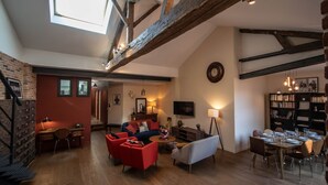 A spacious living room with a high ceiling, exposed wooden beams, a fireplace, a dining area with a table set for six, and a bookshelf filled with books.