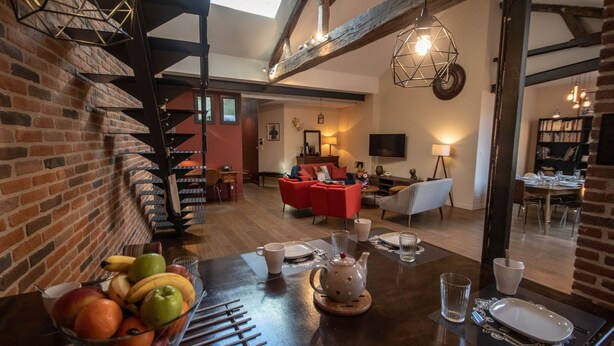 A dining area with a wooden table set for a meal, a fruit bowl, and a cozy living space in the background.