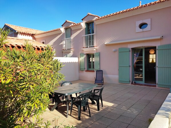 A patio with a table and chairs, a covered seating area, and a building with a tiled roof and green shutters.