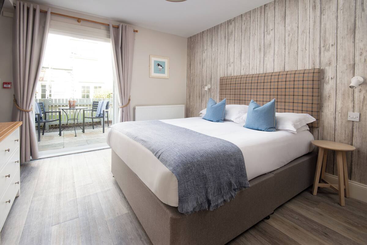 A bedroom with a large bed, a wooden headboard, a small stool, and a view of the outside.