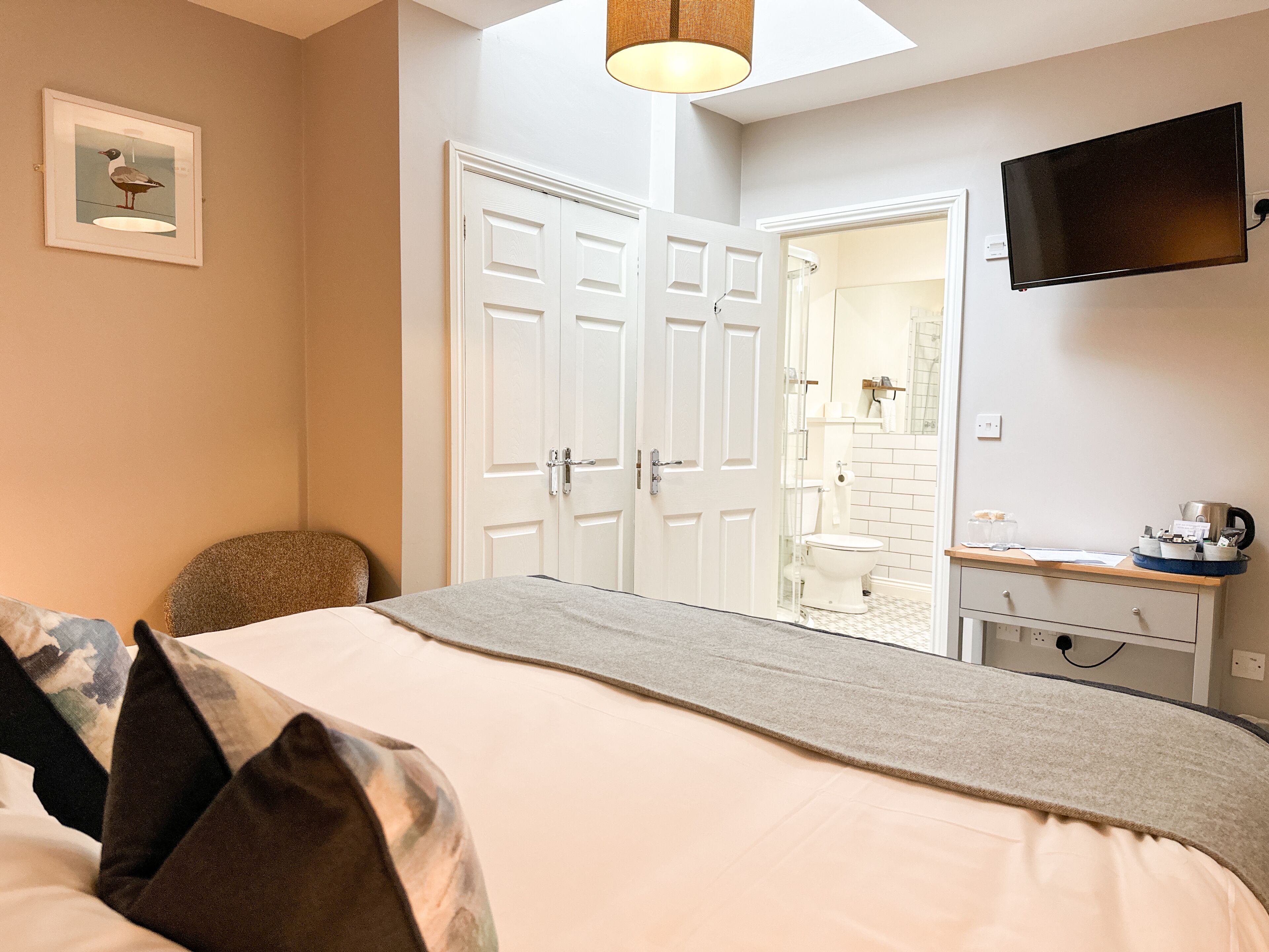 A bedroom with a bed, a chair, a desk with a kettle, and a TV on the wall.