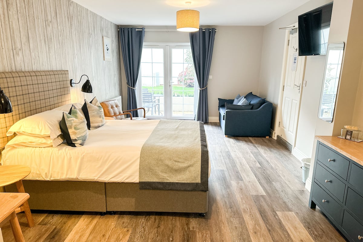 A modern bedroom with a large bed, a sofa, a dining area, and a view of the outdoors.