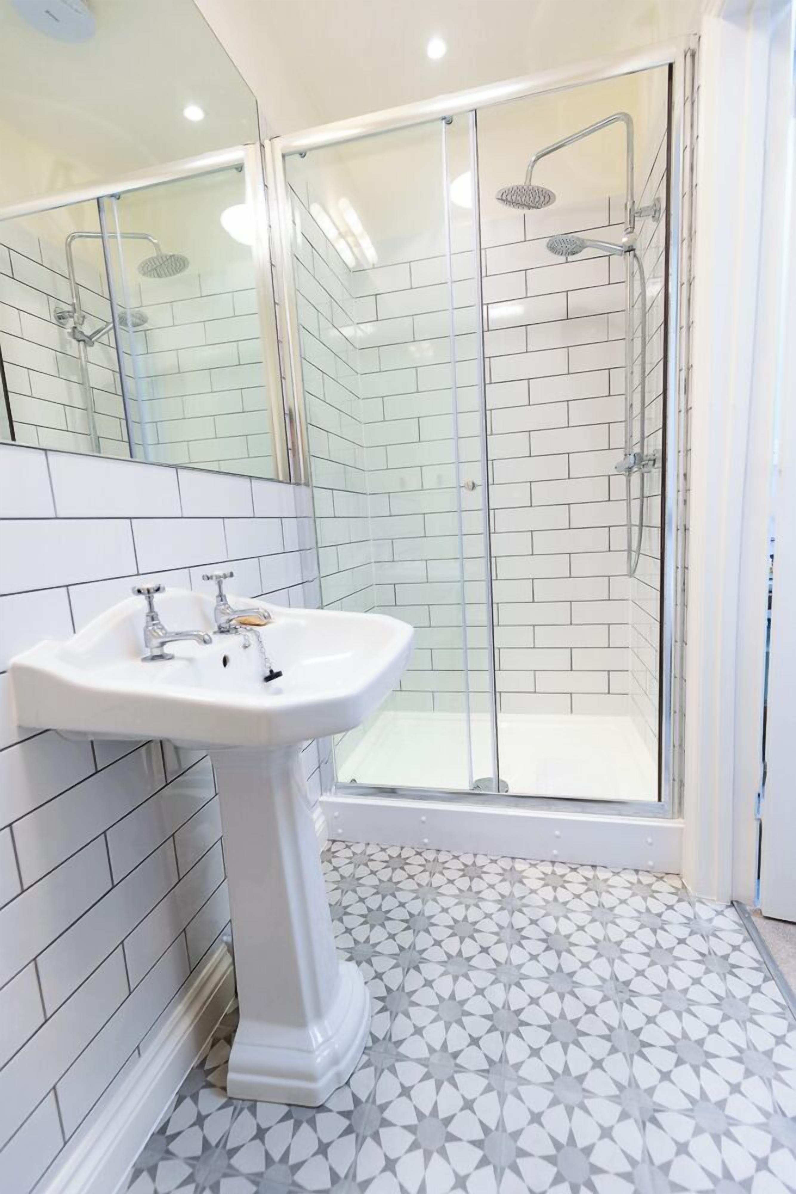 Un baño con un lavamanos blanco de pedestal, cabina de ducha con paredes de vidrio y suelo de baldosas con diseño geométrico.