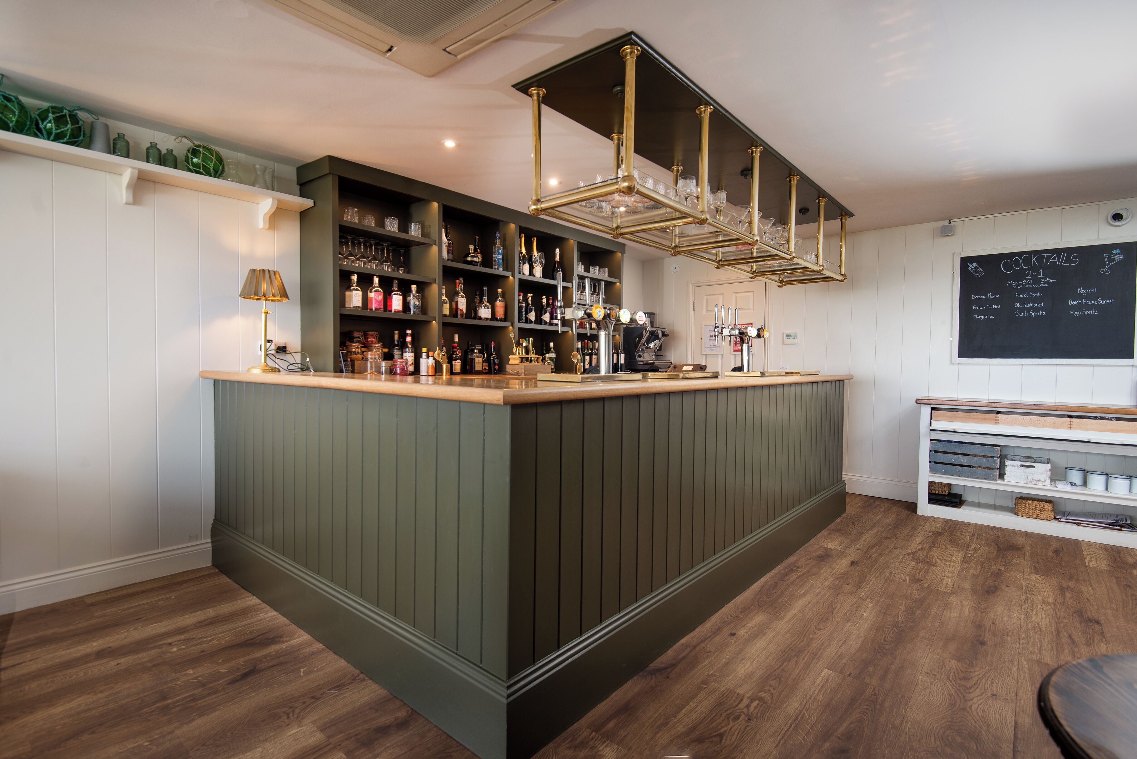 Zona de bar con mostrador de madera, estantes con botellas, un menú en pizarra y una lámpara colgante.