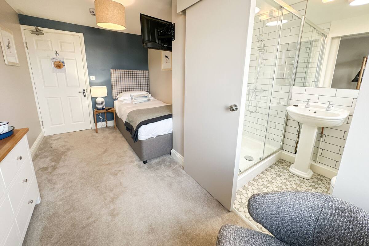 A bedroom with a bed, bedside table, lamp, and a door leading to a bathroom with a shower and sink.