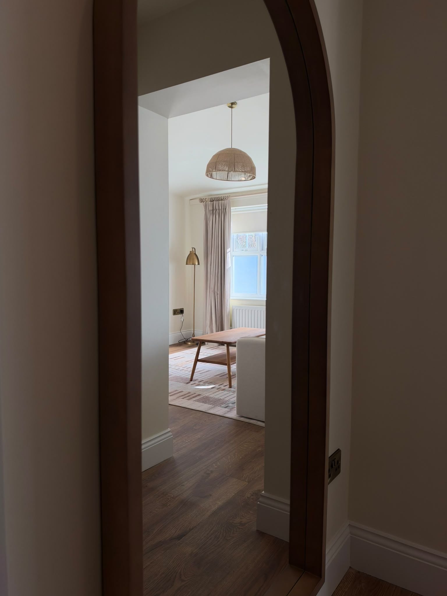 A hallway with a wooden floor, a hanging pendant light, and a view into a room with a window and curtains.