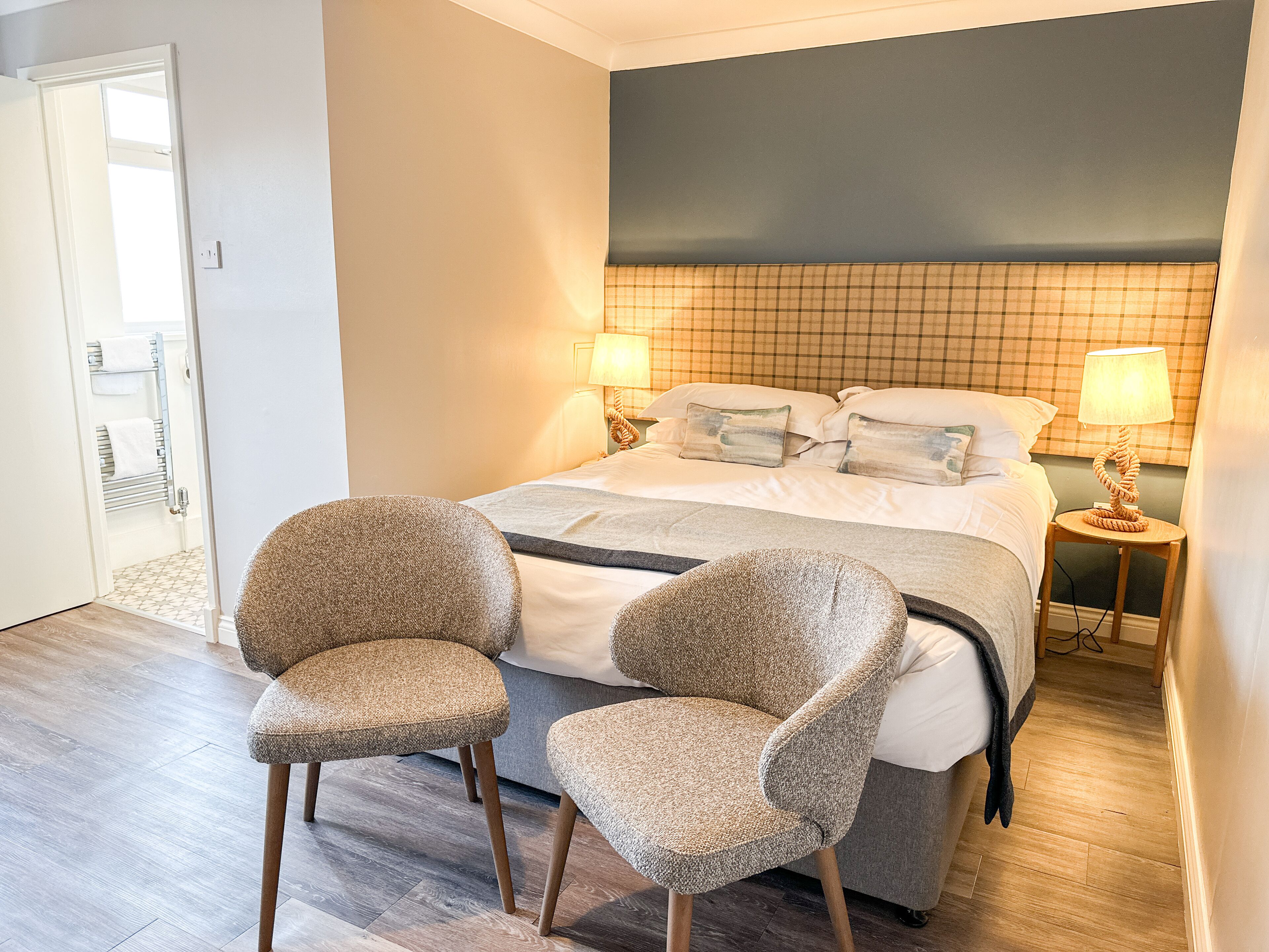A hotel room with a bed, two chairs, a nightstand with a lamp, and a door to the bathroom.