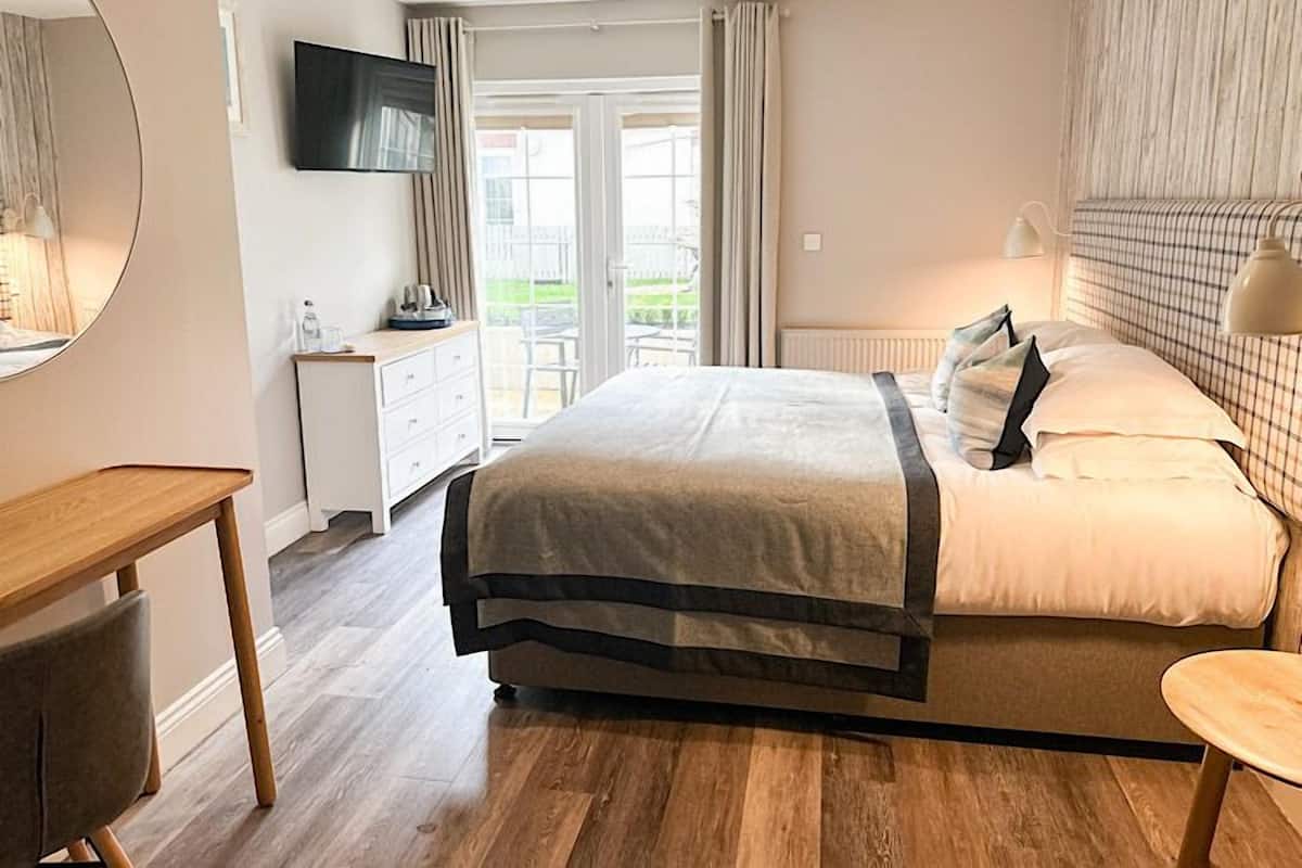 A modern bedroom with a large bed, a bedside table, a dresser, a TV mounted on the wall, and a view of the outdoors through a sliding glass door.