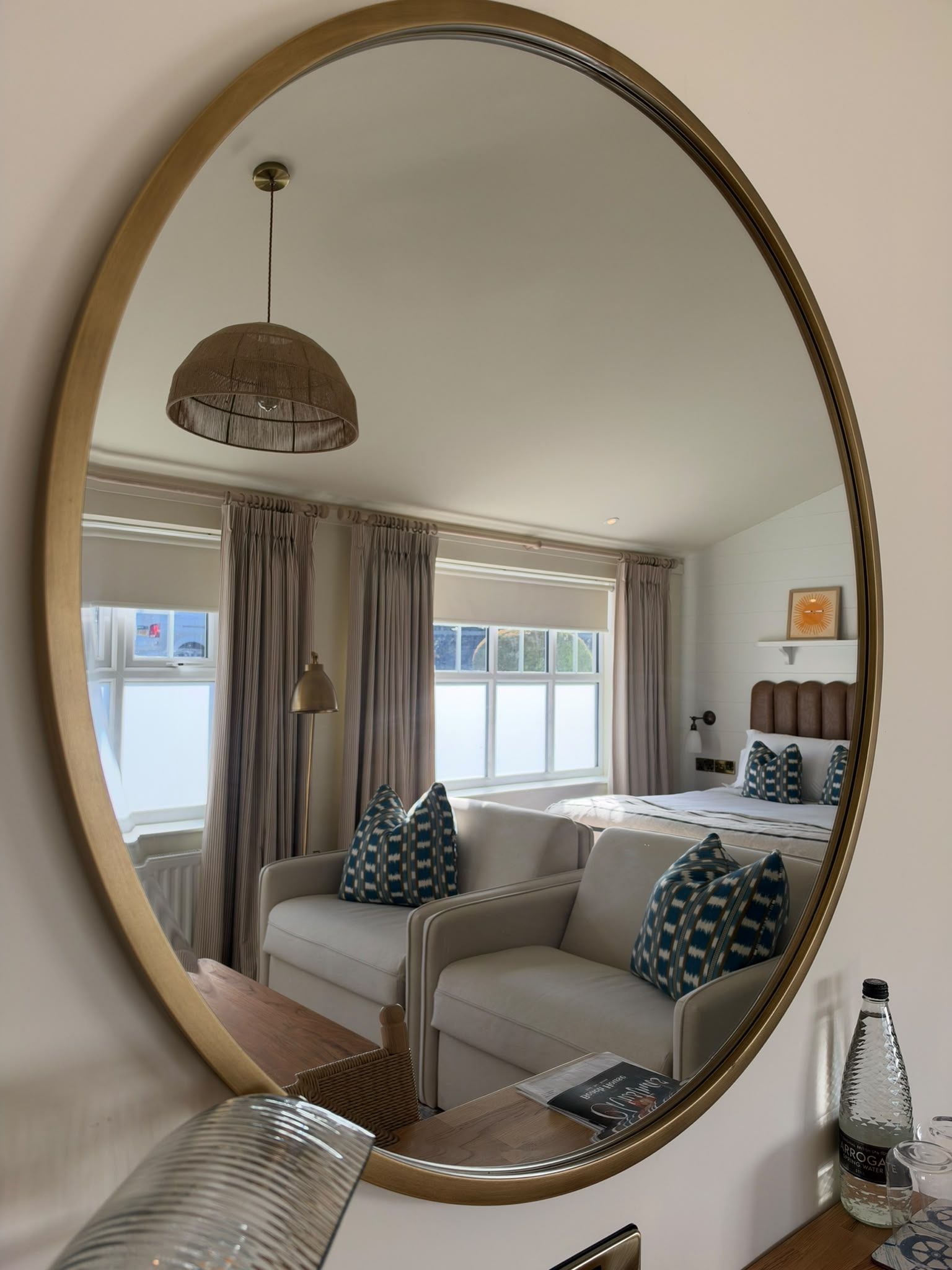 Ein Schlafzimmer mit einem großen, ovalen Spiegel, einem Bett mit gemusterten Kissen, einem Nachttisch mit einer Lampe und einem Fenster mit Vorhängen.