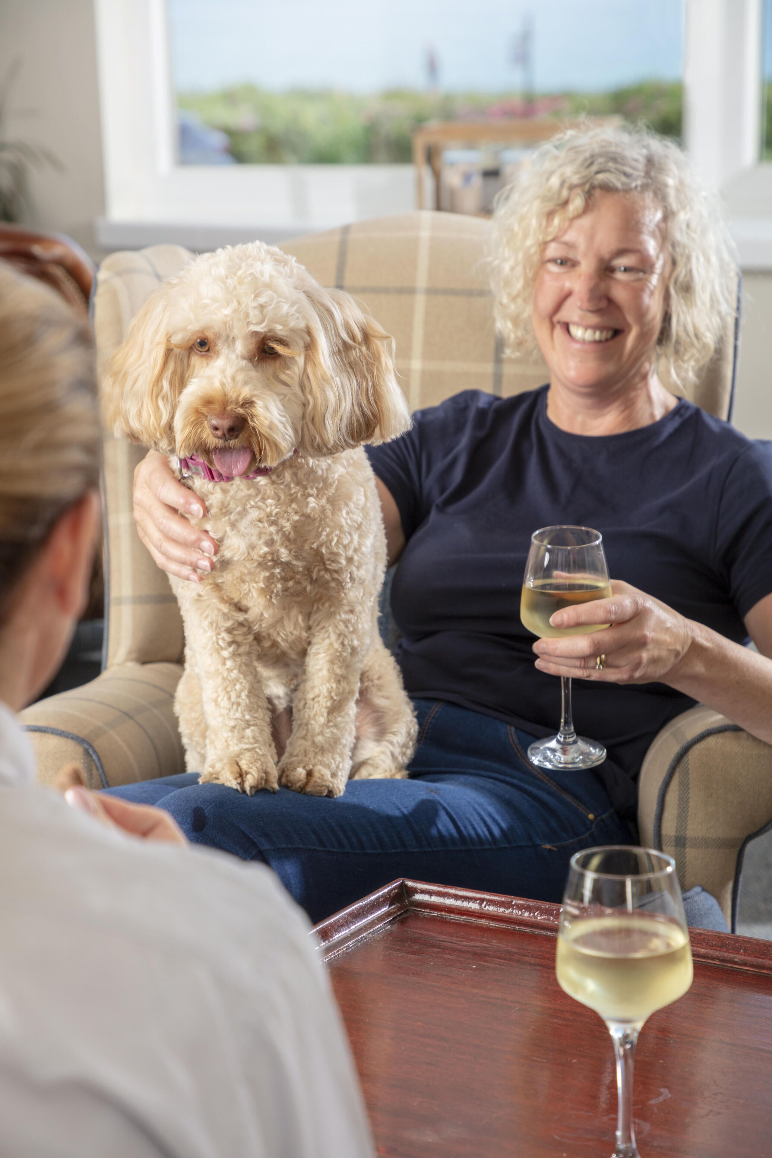 A person sitting on a couch with a dog, holding a glass of wine, and another glass on a table.