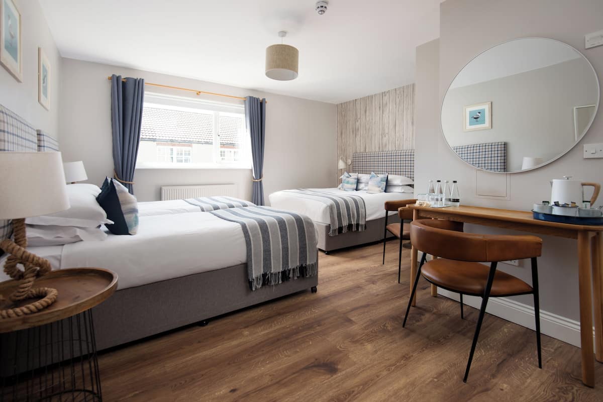 A hotel room with two beds, a desk, a chair, a round mirror, and a window with curtains.