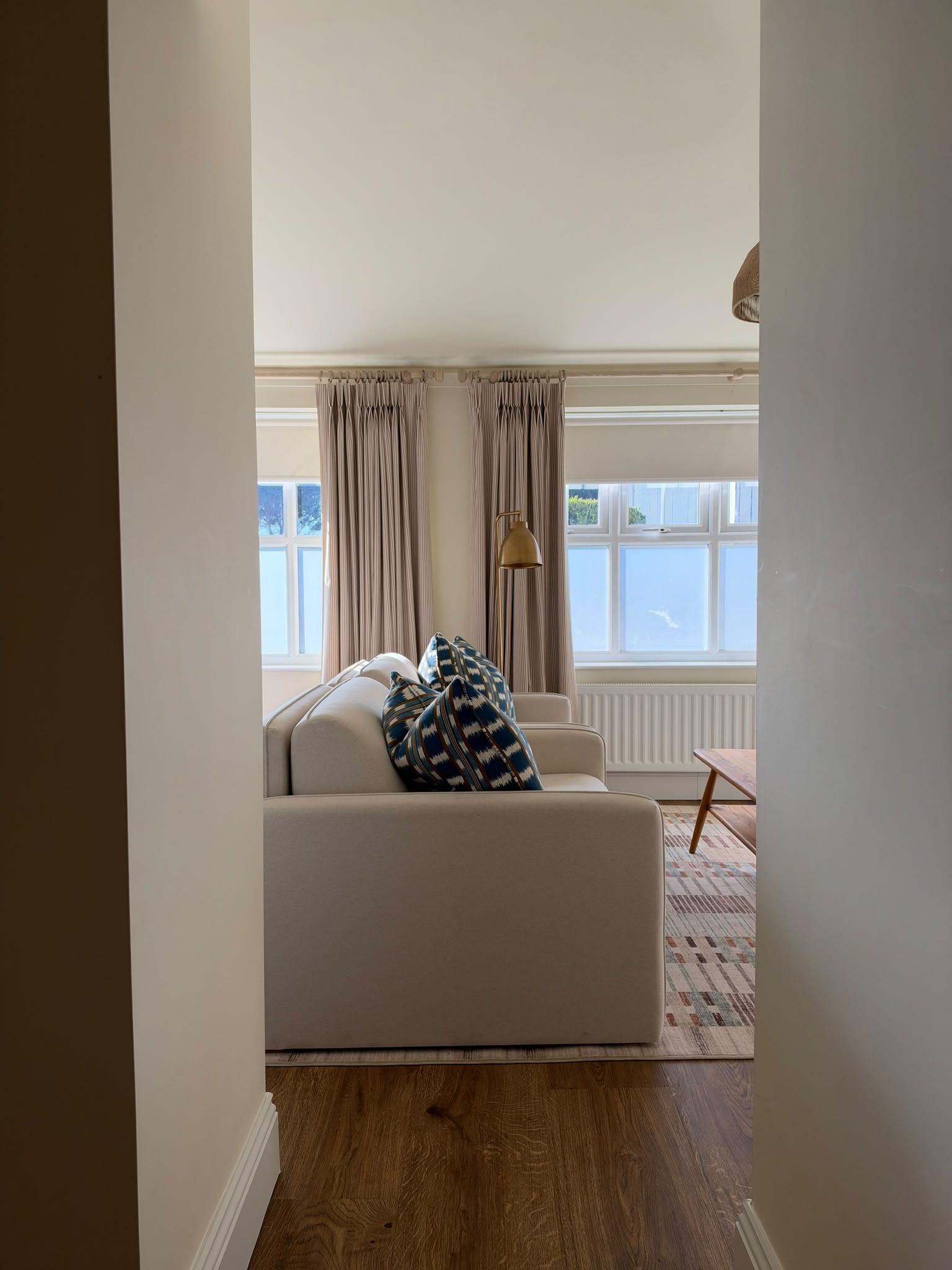 Una habitación con sofá, un cojín estampado, una lámpara y vistas a través de una ventana con cortinas.