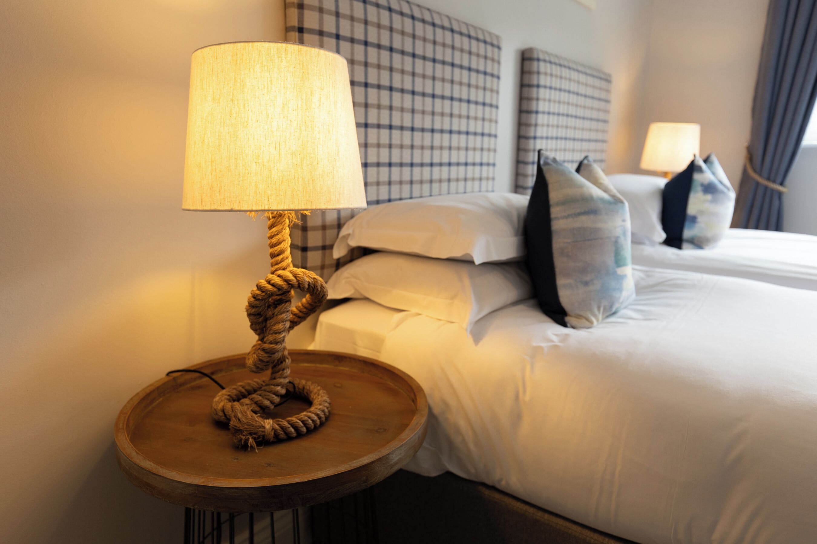 A neatly made bed with white linens and a headboard with a checkered pattern. There is a round wooden nightstand with a lamp and a rope decoration.