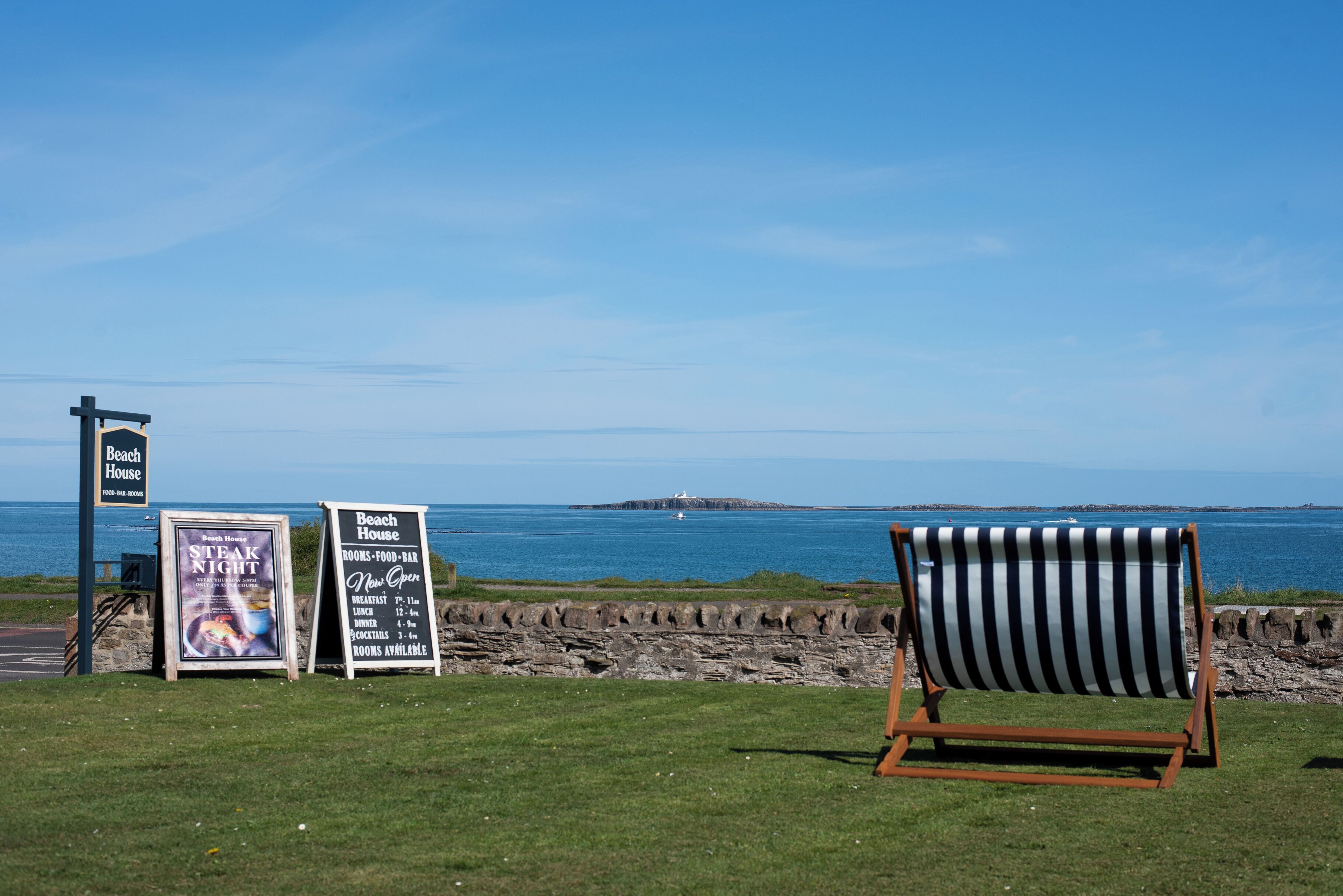 A beach house sign, a poster for a steak night event, and a striped beach chair by the sea.