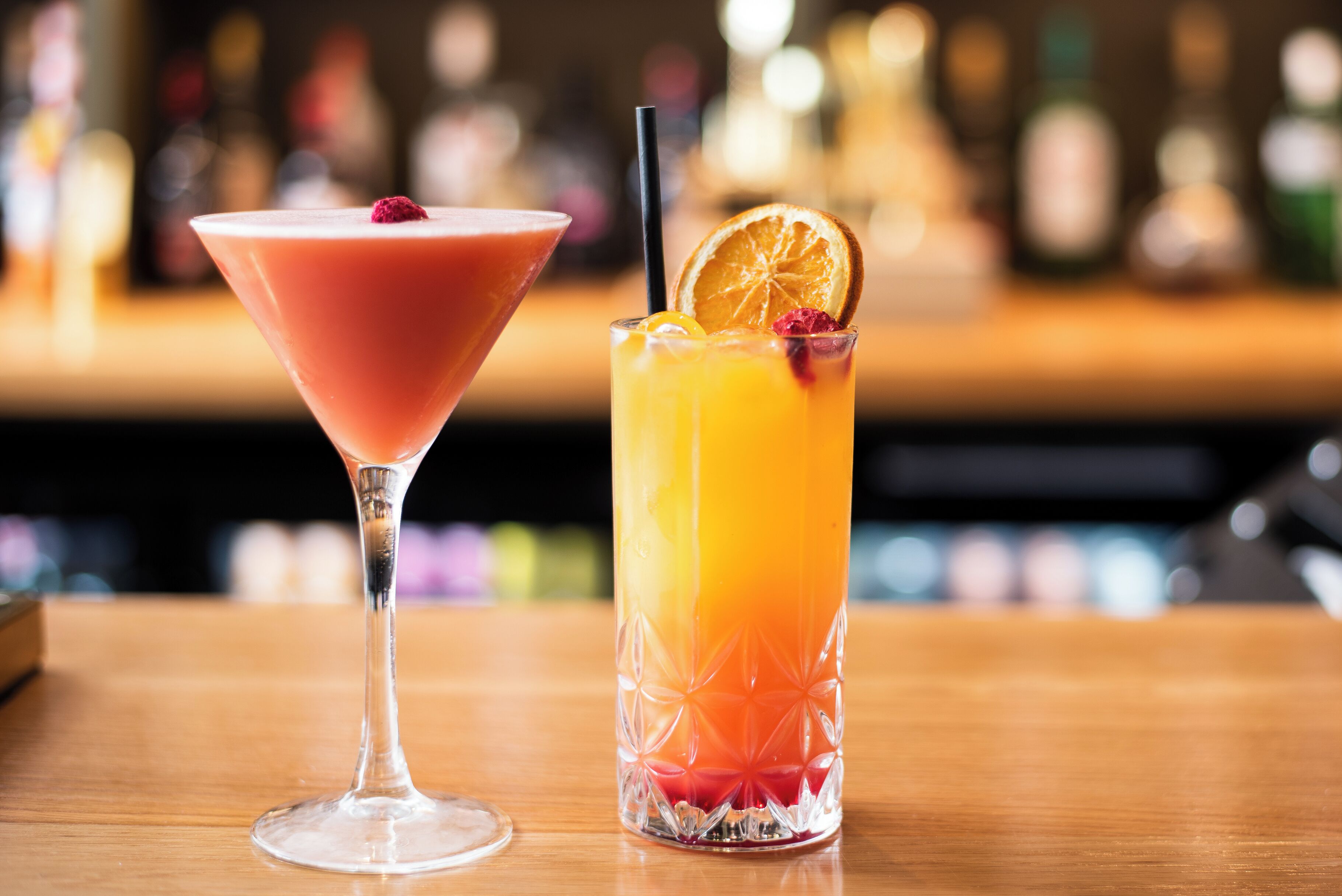 Two cocktails on a bar counter with a blurred background of bottles.