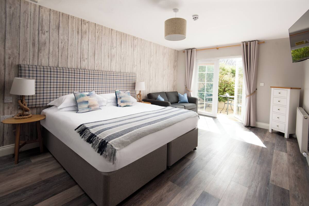 A bedroom with a large bed, a nightstand, a lamp, a sofa, a dresser, and a view of the outdoors.