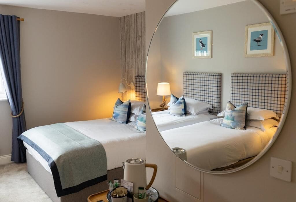 A hotel room with two beds, a large mirror, a window with curtains, and wall-mounted artwork.