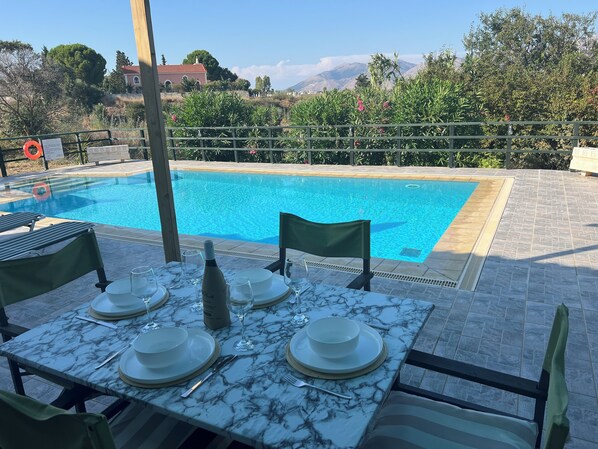 A dining table with plates, glasses, and a bottle, set against a backdrop of a swimming pool and a mountainous landscape. - Villa Varkoula - 2 bedroom family villa with private pool near Xi resort (Mantzavinata)
