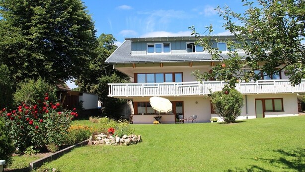 A two-story building with a balcony, a lawn, and a garden with red flowers. - Light-filled, spacious apartment, direct access to terrace and lawn (Ühlingen-Birkendorf)