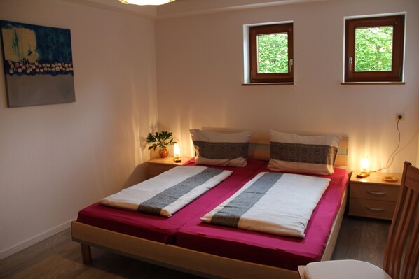 A double bed with a red bedspread and two pillows, a bedside table with a lamp, a small potted plant, and a painting on the wall. - Light-filled, spacious apartment, direct access to terrace and lawn (Ühlingen-Birkendorf)