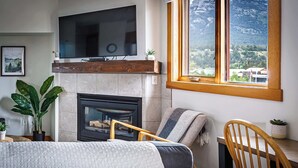 Un salon chaleureux avec une cheminée, un téléviseur fixé au-dessus et une grande fenêtre donnant sur une vue imprenable sur les montagnes.